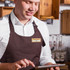 A wooden name badge is worn on a shirt, with a brown apron and a person using a tablet in a café setting.