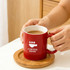 A red ceramic mug with a white interior, featuring a logo and sitting on a wooden coaster.