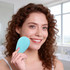A silicone facial cleansing brush in light blue held by a smiling person with curly hair in a bright bathroom setting.