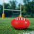 A red mini promotional Aussie Rules ball resting on green grass, with goalposts blurred in the background.