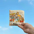 A foldable shopping bag in soft yellow with colourful cartoon characters and a logo, set against a blue sky.