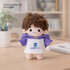 A plush doll with brown hair wearing a purple raglan hoodie and a white front, featuring a logo.