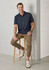A man sits on a wooden stool, wearing a navy short sleeve polo and light brown pants with white shoes. The polo has a logo.