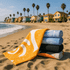 A stack of five bath towels in various colours, including yellow, blue, and black, on a sandy beach with palm trees and houses in the background.