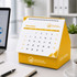 A yellow desk calendar with an organiser, featuring a spiral binding and displayed on a workspace with a notebook and laptop.