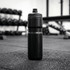 A drink bottle in black, standing upright on a gym floor with weights in the background.