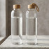 A glass drink bottle with a wooden lid and a rope loop, containing clear water, featuring a sleek design.