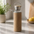A drink bottle featuring a glass body, a metal lid, and a tan fabric sleeve, set on a kitchen countertop.