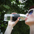 A drink bottle with a clear body and a black silicone grip, held by a person sipping water outdoors.