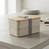 A two-tiered lunch box in neutral tones with a wooden lid and a grey strap, resting on a smooth table.