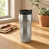 A sleek silver travel mug with a black lid and a logo, placed on a wooden table next to a plant and notebook.