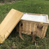 A square wicker picnic cooler basket in natural tones with a bamboo lid and brown straps, placed on grass.
