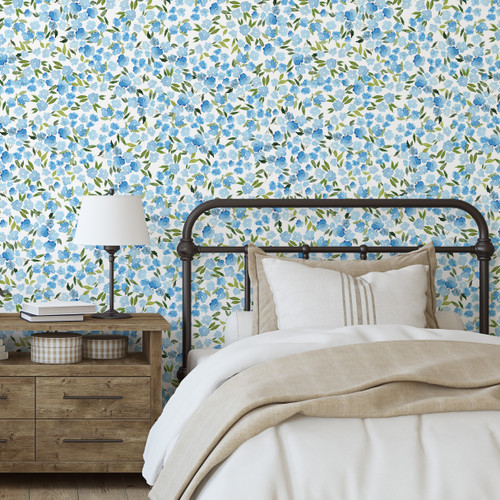 A farmhouse style bedroom with a black metal bed, a wooden side table with a lamp and watercolor forget me not flowers on the wallpaper