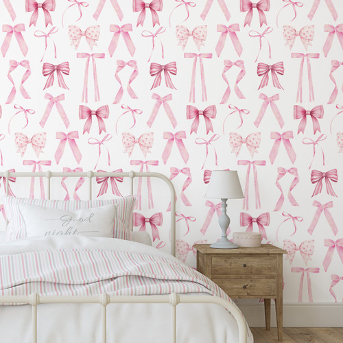 A bedroom with white and pink bow-patterned wallpaper. A white metal bedframe holds a bed adorned with a striped pink and white comforter and pillows, including one with the phrase "sweet wishes." A wooden nightstand with a white lamp sits beside the bed.