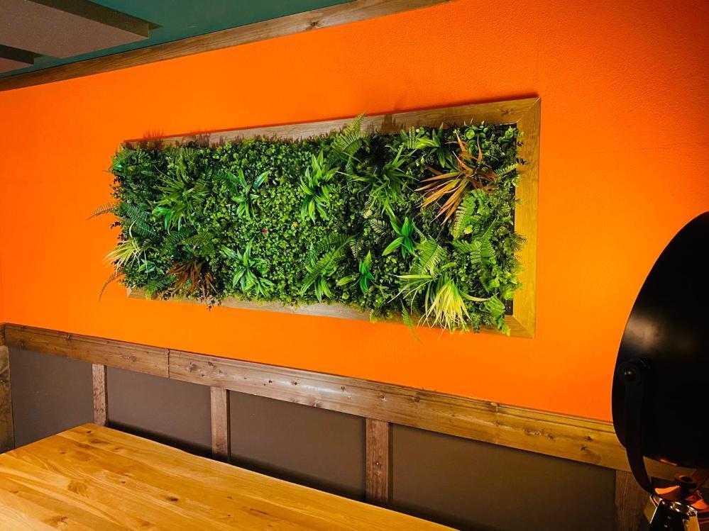 DIY artificial green wall panel installed on orange wall showing lush foliage in wooden frame