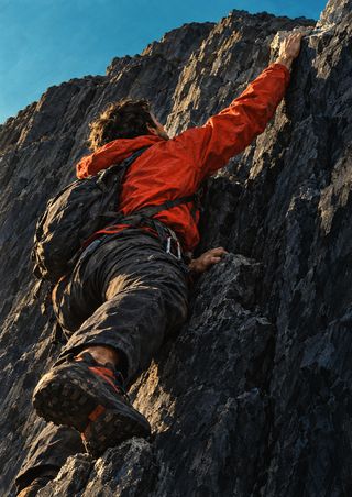 Climber in a vermilion-orange jacket reaching up a dark ridged rock face with a small area of turquoise sky above.