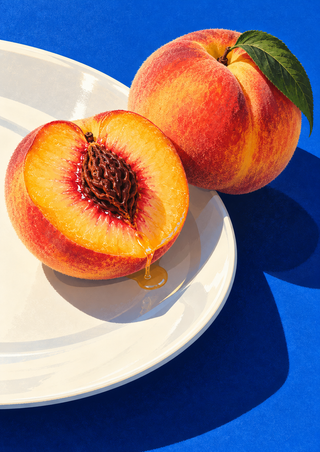 Cut peach with exposed pit and whole peach with a green leaf on a white plate against a cobalt-blue surface.