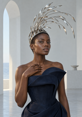 A fashion model wears a navy strapless gown and a tall silver reed-like headdress in a pale interior with an arched sea view.