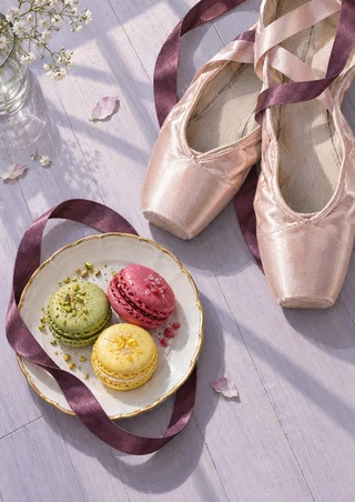 Pale pink ballet slippers with plum ribbons beside three colorful macarons on a gold-rimmed plate over a pale lilac floor.