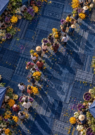 Overhead view of shoppers and vendors moving among yellow and purple flowers, baskets, and patterned indigo mats.