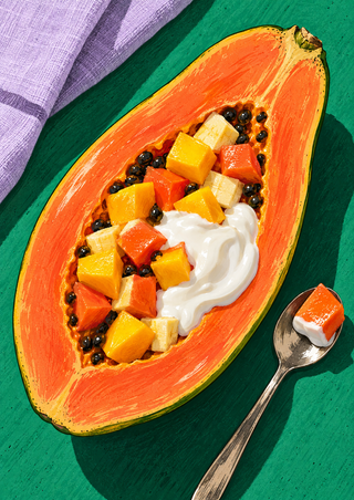 Halved papaya filled with colorful fruit cubes, dark seeds, and yogurt beside a spoon on a jade-green surface.