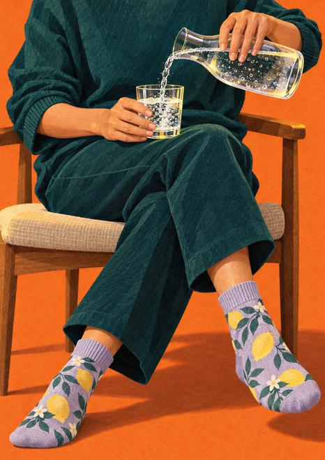 Faceless seated person in dark teal clothing pours sparkling water into a tumbler beside crossed legs in patterned lavender socks against orange.