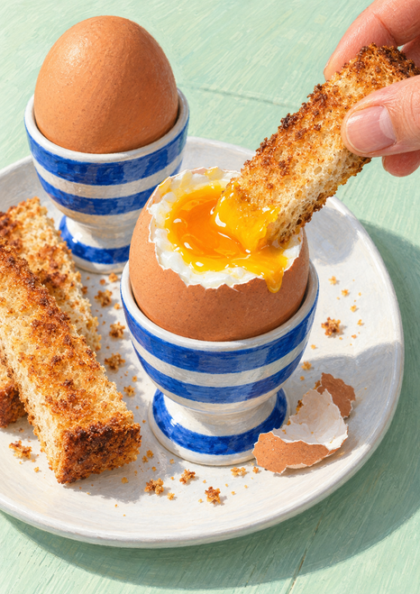Hand dipping toast into a cracked soft-boiled egg in a blue-and-white striped cup, with a second egg and extra toast on a plate.