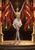 Grey-skinned dark elf in gold and white lifting red banners on a temple stage before an empty auditorium.