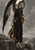 Lion-headed figure in a black robe looks back from stone steps, with a large feathered wing and distant ruins.