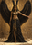 Full-length winged woman in Egyptian-inspired jewelry and a dark feathered skirt before a gold-toned stone wall with carved-looking motifs.