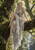 Silver-haired pointed-eared woman in a pale leaf-patterned dress sitting on a mossy branch with a waterfall in the forest.