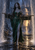 Gray-skinned dark-haired elf in an emerald gown holds a horizontal spider-decorated standard between two curtains of falling water.