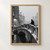 Bundled pedestrian on a snow-covered stone footbridge over dark canal water, with a lit arch and snowy city beyond. in a styling frame; frame not included