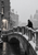 Bundled pedestrian on a snow-covered stone footbridge over dark canal water, with a lit arch and snowy city beyond.. Complete unframed artwork at full portrait size
