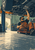 A mechanic on a wheeled creeper reaches beneath a raised vehicle in a garage with tools and shelving.