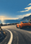 Unbranded orange sports car on a sweeping coastal road beside a stone breakwater, sea and distant rocky headland.