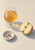Stemmed glass of amber drink beside a halved apple and a shallow dish of seeds on a pale tabletop.