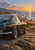 A deep-green classic coupe sits on rocky coastal ground, with the sea, distant headland, surf, and low golden sun behind it.