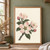 Pink apple blossoms and buds on a diagonal woody branch with dark leaves against a warm ivory background.; frame shown for styling only