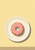 Overhead still life of a pink-glazed, sprinkled doughnut centered on a cream plate against a pale yellow ground.