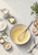 Overhead kitchen work surface with a bowl of pale yellow mixture, whisk, ingredients, herbs, utensils, and folded cloth.