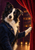 Black-and-white border-collie-like dog in a dark blue jacket touching a red theater curtain, with warm auditorium lights behind it.