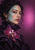 Adult woman with dark wavy hair and a turquoise branching light pattern over one cheek and temple, wearing a dark violet garment with a large purple orchid against a magenta-violet background.