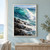 Turquoise breaking wave with white spray and foam above dark foreground rocks, beneath a pale sky and distant horizon.; frame shown for styling only