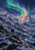 Two hikers on a snowy ridge look toward a sweeping green, teal and violet aurora above snowy mountains and reflective valley water.