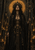 Crowned woman in black-and-gold robes holds a circular-topped staff in a candlelit stone interior.