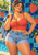 Confident woman in a coral top and denim shorts poses with a denim jacket before a strawberry-decorated poolside wall.
