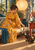 A woman in a golden-saffron robe reaches to play with a tortoiseshell cat in a sunroom with a paper lantern, floral curtains, plants, and a red table.