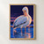 Large white pelican preening in shallow electric-blue water beneath a dark violet sky with orange reflections.; frame shown for styling only
