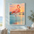 Coral-pink flamingo standing on one leg in shallow teal water, with its other leg raised beneath a peach sunset sky.; frame shown for styling only
